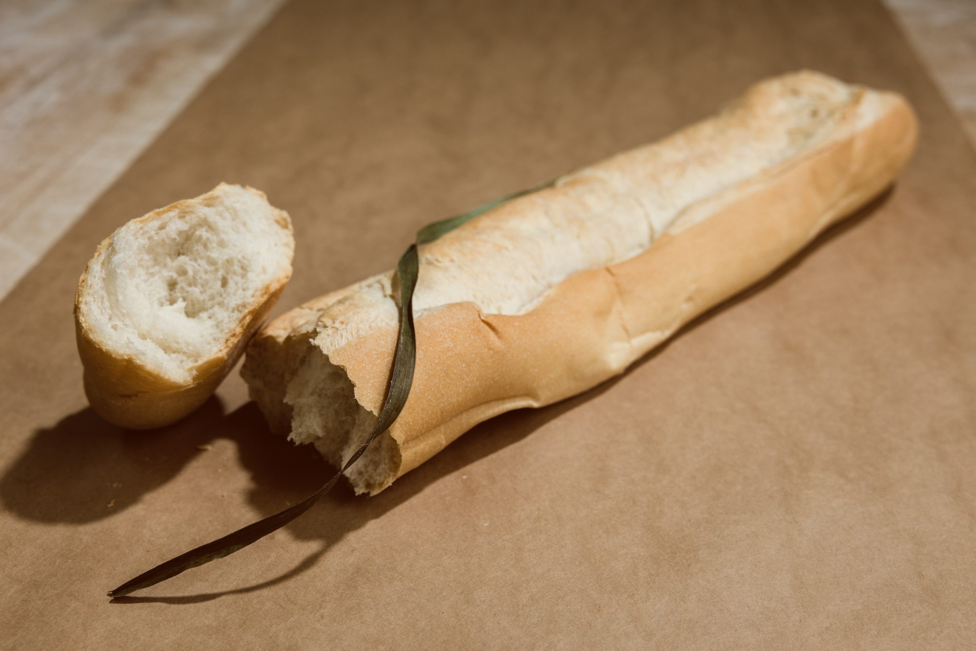 An 18-inch La Segunda Cuban bread loaf laid horizontally on craft paper, one end torn open to reveal the airy interior crumb, palmetto leaf still resting on top.